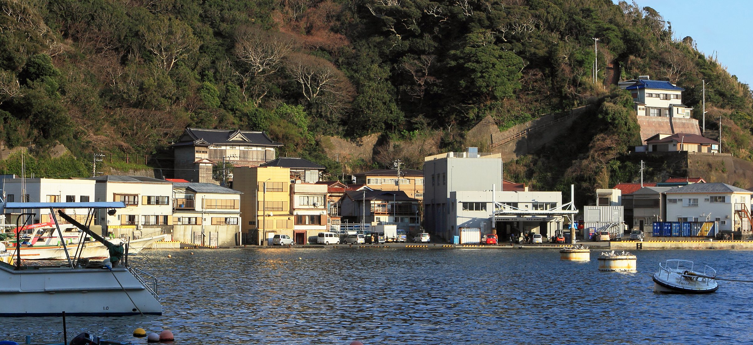 A small Japanese harbour town at dusk, houses stacked on the hillside above the boats